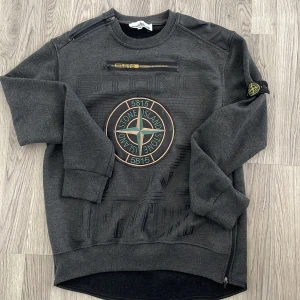 Grå sweatshirt från Stone Island - Säljer en snygg grå sweatshirt från Stone Island med deras ikoniska logga på bröstet. Tröjan är tillverkad i 100% bomull och har långa ärmar samt ribbade muddar. Perfekt för en stilren look.