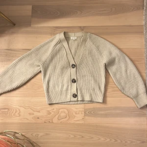 Beige stickad kofta från H&M - Säljer en mysig beige stickad kofta från H&M. Koftan har en v-ringad design med stora knappar framtill och ribbade detaljer. Perfekt för kyliga dagar!