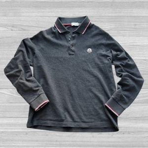 Moncler pikétröja - Säljer en stilren grå pikétröja från Moncler med långa ärmar och klassisk krage. Tröjan har röda och vita detaljer på kragen och ärmsluten samt en broderad logotyp på bröstet. Perfekt för en avslappnad men snygg look. Storleken är M men passar bättre till S. (Priset kan diskuteras❗️)