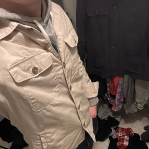 Beige jacka med knappar - Säljer en stilren beige jacka med knappar och två bröstfickor med lock. Jackan har en klassisk krage och är långärmad, perfekt för vår och höst. Passar bra över en hoodie för en avslappnad look. Skriv om du undrar något om plagget!🙌😊