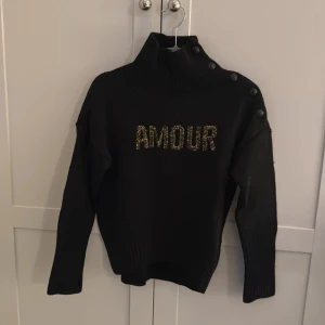 Svart tröja med text från Zadig & Voltaire - Snygg svart tröja från Zadig & Voltaire med texten 'AMOUR' i glittriga bokstäver på framsidan. Tröjan har en hög krage och knappar längs axeln. Den här tröjan har inte används så mycket tröjan kommer även med en extra knapp på lappen inuti tröjan