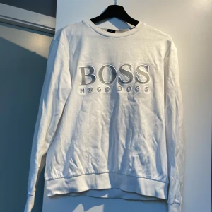 Vit tröja från Hugo Boss - Säljer en stilren vit tröja från Hugo Boss med stort BOSS-tryck på framsidan. Tröjan har en slim fit passform och långa ärmar. Perfekt för en avslappnad men ändå snygg look. Det finns två små fläckar en som är svart målarfärg på höger armen och en liten prick men det är inget som man tänker på när man har på sig den