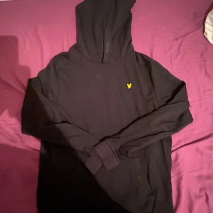 Svart hoodie från Lyle & Scott - Säljer en svart hoodie från Lyle & Scott med en gul logga på bröstet. Tröjan har en klassisk design med huva och långa ärmar. Perfekt för en avslappnad stil.