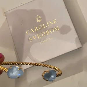 Guldarmband med blå stenar från Caroline Svedbom - Helt nytt guldarmband elegant  från Caroline Svedbom med två ljusblå stenar. Armbandet har en tvinnad design som ger en lyxig känsla. Jag säljer armbandet för jag har ett till och då kommer detta aldrig till användning 