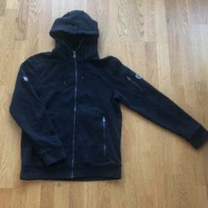 Svart hoodie från Nautic XPRNC - Säljer en svart hoodie från Nautic XPRNC med dragkedja och justerbar huva. Tröjan har långa ärmar och broderad logga på bröstet. Perfekt för en avslappnad stil.