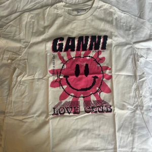 Vit t-shirt från Ganni med coolt rosa tryck - Säljer en vit t-shirt från Ganni med ett stort rosa smiley-tryck och texten 'LOVE CLUB'. Perfekt för en avslappnad stil. T-shirten har korta ärmar och en rund halsringning med en oversized fit. Perfekt till sommaren! Passar s-m💞💞💞