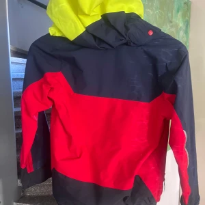 Flerfärgad vindjacka från Helly Hansen - Säljer en cool flerfärgad vindjacka från Helly Hansen. Jackan har en kombination av mörkblått, rött och neongult med dragkedja framtill. Perfekt för blåsiga dagar med sin skyddande huva. Namnad