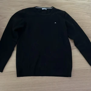 Svart kofta från J.Lindberg  - Snygg svart kofta från J.Lindberg med en liten vit logga på bröstet. Tröjan har en klassisk design med långa ärmar och rund halsringning. Perfekt för en stilren look.