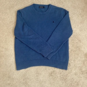 Blå tröja från Polo Ralph Lauren - Säljer en snygg blå tröja från Polo Ralph Lauren med rund hals och ribbade muddar. Den har en diskret broderad logga på bröstet. Perfekt för en stilren look.