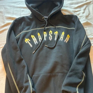 Svart hoodie från Trapstar - Säljer en svart hoodie från Trapstar med gul och vit text på bröstet. Hoodien har en klassisk design med dragsko i huvan och en stor magficka. Perfekt för en avslappnad stil.