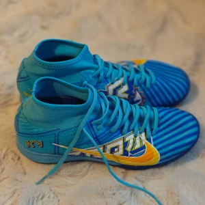 Blå och gula Nike fotbollsskor - Snygga blå fotbollsskor från Nike med gula detaljer och randigt mönster. Skorna har snörning och är designade för inomhusbruk med en slitstark sula. Perfekta för den som vill sticka ut på planen. Mbappe 