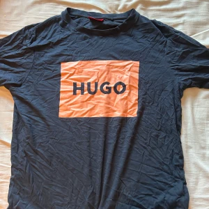 Svart t-shirt från Hugo - Säljer en svart t-shirt från Hugo med en stor orange logga på framsidan. T-shirten är kortärmad och har en klassisk rund halsringning. Perfekt för en avslappnad stil.