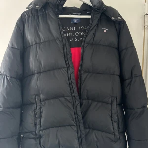 Svart dunjacka från GANT - Snygg svart dunjacka från GANT med huva och dragkedja. Jackan har en klassisk design och är väldigt varm och skön. 