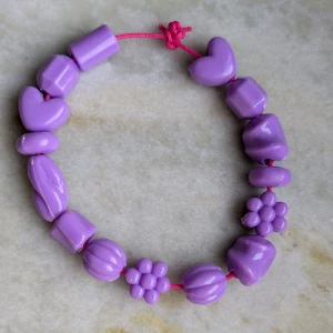 Lila armband med hjärtan och blommor - Säljer ett sött lila armband med olika former som hjärtan och blommor. Armbandet är trätt på ett rosa snöre och har en lekfull design. Perfekt för att lägga till en färgglad touch till din stil!