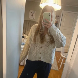 Beige stickad kofta  - Mysig beige stickad kofta med kabelstickat mönster och knappar framtill. Perfekt för kyliga dagar med sin långa ärmar och v-ringade design. En klassisk och stilren kofta som passar till det mesta.