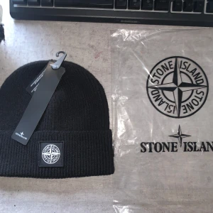 Svart stickad mössa från Stone Island - Snygg svart stickad mössa från Stone Island med deras ikoniska logga framtill. Perfekt för att hålla värmen under kyliga dagar. Mössan har en ribbad design och kommer med originalförpackning.
