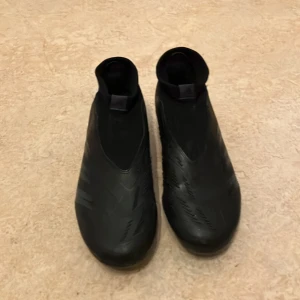Svarta fotbollsskor från Adidas - Snygga svarta fotbollsskor från Adidas med en stilren design och diskreta ränder. Skorna har en tight passform och är perfekta för spel på gräs. De har en låg profil och är tillverkade i syntetmaterial för optimal prestanda.