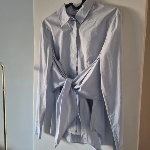Ljusblå skjorta med knyt från H&M - Snygg ljusblå skjorta från H&M med långa ärmar och en unik knytning framtill. Perfekt för en stilren look. Skjortan har en klassisk krage och är tillverkad i ett lätt och bekvämt material.