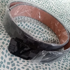 Grått bälte med svart spänne - Snyggt grått bälte med ett stilrent svart spänne. Perfekt för att ge en elegant touch till din outfit. Bältet har en texturerad yta och är inslaget i plast för skydd.
