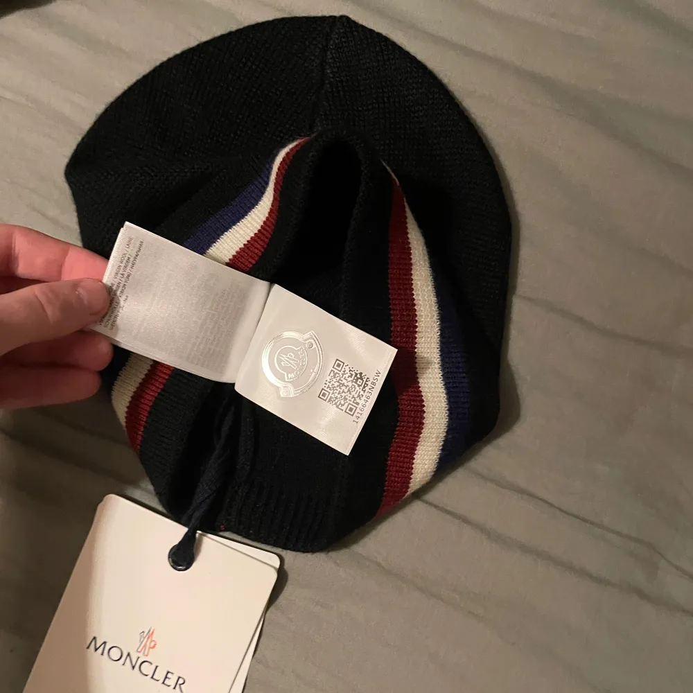 Snygg svart mössa från Moncler med ränder i rött, vitt och blått. Den har en broderad logotyp framtill och är perfekt för kyliga dagar. Stilren och enkel att matcha med olika outfits. Aldrig använd . Asusteet.