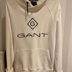 Vit hoodie från GANT - Säljer en stilren vit hoodie från GANT med svart logotyp på bröstet. Tröjan har en klassisk känguruficka och justerbar huva med snören. Perfekt för en avslappnad look.