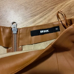 Brun kjol från BikBok - Snygg brun kjol i skinnimitation från BikBok. Kjolen har en dragkedja baktill och är i en kort modell. Perfekt för att ge en stilren look.
