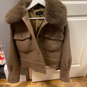 Meotine jacka - Snygg brun jacka från Meotine med en lyxig pälskrage. Jackan har knappar framtill och två stora fickor med lock. Perfekt för kyligare dagar med sin stilrena design och varma material. Den är i bra skick 