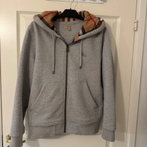 Grå hoodie från Burberry - Säljer en stilren grå hoodie från Burberry med klassiskt rutigt mönster i huvan. Tröjan har dragkedja och snörning vid huvan samt praktiska fickor framtill. Perfekt för en avslappnad och trendig look.