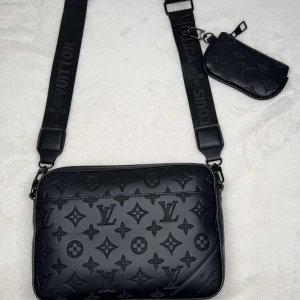 Svart axelväska med monogram - Snygg svart axelväska med monogrammönster och justerbar axelrem. Väskan har en medföljande liten plånbok som kan fästas på remmen. Perfekt för att bära med sig det viktigaste med stil.