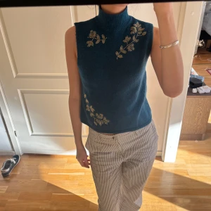 Blå ärmlös polotröja med broderi och pärlor - Snygg blå ärmlös polotröja med vackra handbroderade blommor med guldiga pärlor. Perfekt för en stilren look med en twist. Polokragen ger en elegant touch.