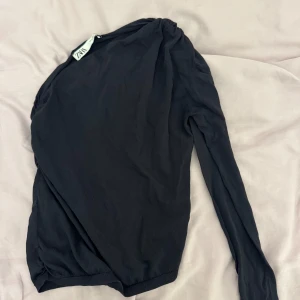 Svart långärmad topp från Zara - Snygg svart långärmad topp från Zara med en elegant drapering över axeln. Perfekt för en stilren look. Materialet känns mjukt och bekvämt mot huden.
