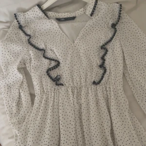 Vit prickig klänning från Zara - Säljer en söt vit klänning från Zara med små svarta prickar och volangdetaljer. Klänningen har långa ärmar och en v-ringad design. Perfekt för en somrig look!