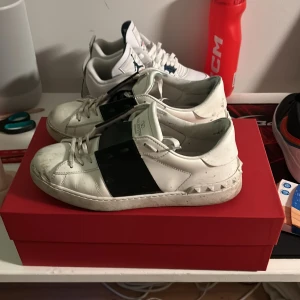 Valentino Garavani sneakers i vitt och svart - Snygga Valentino Garavani sneakers i vitt med svarta detaljer. Skorna har en unik design med nitar runt sulan och snörning framtill. Perfekta för att ge din outfit en lyxig touch.