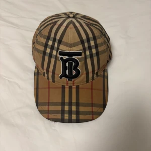 Äkta Burberry keps - Äkta där kvitto finns från NK. Varan är köpt flera år sedan, användes mycket då men nu har den bara legat i garderoben i cirka 2-3 år oanvänd. Kepsen har även ett litet snitt fram på skärmen som går att se lite på första bilden. 