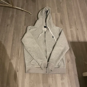 Grå hoodie från Ralph Lauren - Säljer en stilren grå hoodie från Ralph Lauren med dragkedja och klassisk logga på bröstet. Perfekt för en avslappnad look. Den har en justerbar huva med snören och ribbade muddar vid ärmslut och nederkant. Det går fortforande att anvönda dragkedjan men själva saken man drar på är borta