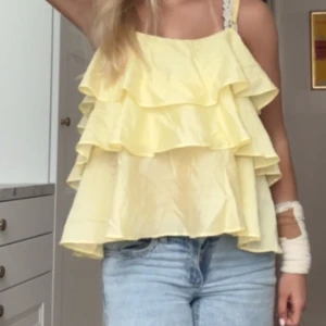Gul volangtopp - Säljer en söt gul topp med volanger och smala axelband. Perfekt för sommardagar och ger en luftig och lekfull look. Passar bra till jeans eller kjol för en avslappnad stil.