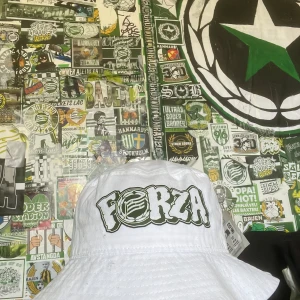 HAMMARBY BUCKET HATT! - Snygg vit bucket hat från Atlantis med texten 'Forza' i grönt och svart tryck. Perfekt för att visa ditt stöd på matchen eller bara som en stilren accessoar. Tillverkad i bomull för en bekväm passform.