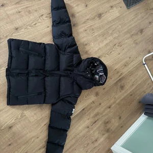 Burberry jacka/väst  - Säljer en snygg svart pufferjacka från Burberry med huva och dragkedja. Jackan har ett klassiskt rutigt mönster på insidan av huvan och Burberry-logga på ärmen. Perfekt för kyliga dagar! Aldrig använd