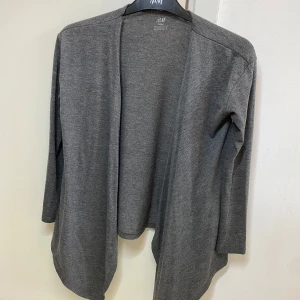 Grå kofta från H&M - Säljer en stilren grå kofta från H&M. Den har en öppen design utan knappar och långa ärmar, perfekt för lager på lager-stil. Passar bra till både vardag och mer uppklädda tillfällen.