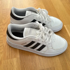Vita och svarta sneakers från Adidas - Snygga vita sneakers från Adidas med klassiska svarta ränder på sidorna. Skorna har snörning och en bekväm sula med bra grepp. Perfekta för en avslappnad stil.