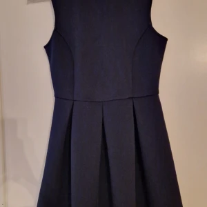 Mörkblå klänning från Nelly - Elegant mörkblå klänning från NLY One med ärmlös design och veckad kjol. Klänningen har en stilren öppen ryggdetalj som ger en extra touch. Perfekt för en kväll ute eller en speciell tillställning.