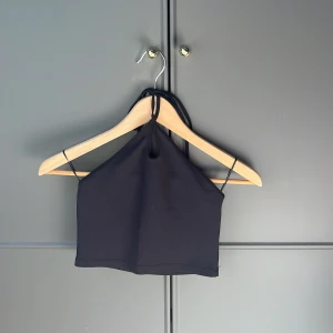 Svart halterneck topp - Snygg svart halterneck topp med ribbad textur. Toppen har en stilren design med smala axelband och en liten öppning framtill. Perfekt för en trendig look.