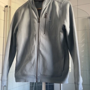 Ljusblå zip tröja från Sail Racing - Säljer en stilren ljusblå zip tröja från Sail Racing med dragkedja framtill och en praktisk bröstficka med dragkedja. Perfekt för en sportig look. Tröjan har långa ärmar och en bekväm passform.