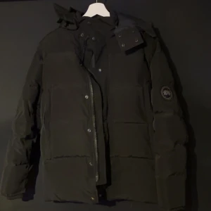 Svart pufferjacka med huva - Säljer en stilren svart pufferjacka med huva och dragkedja. Jackan har långa ärmar och en diskret logga på ärmen. Perfekt för kyliga dagar.