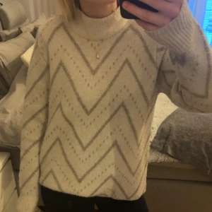 Beige stickad tröja med mönster - Säljer en mysig beige stickad tröja med ett snyggt zickzackmönster i grått glitter. Tröjan har en hög krage och långa ärmar, perfekt för kyliga dagar. Den är både stilren och bekväm. Nästan aldrig använd.