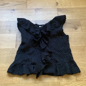Svart topp från Gina Tricot - Säljer en svart topp från Gina Tricot med volangdetaljer. Toppen har en unik struktur och är ärmlös. Den har en vacker design med volanger längs framsidan och nederkanten. Köpt från Gina tricot i storlek 146/152, knappt använd