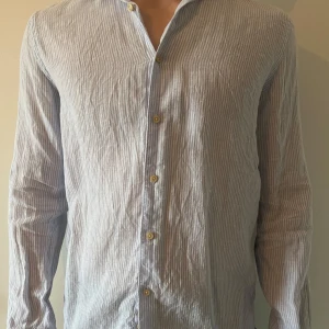 Randig skjorta med knappar - Snygg och stilren långärmad skjorta med smala ränder i ljusblått och vitt. Skjortan har en klassisk krage och knappar framtill. Perfekt för en avslappnad men ändå uppklädd look.