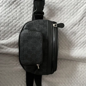 Svart axelväska med mönster - Snygg svart axelväska med diskret mönster och justerbar axelrem. Väskan har flera dragkedjeförsedda fack för praktisk förvaring. Perfekt för att hålla dina saker organiserade och lättillgängliga.