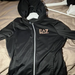 Svart jacka från Emporio Armani - Snygg svart jacka från Emporio Armani med dragkedja och huva. Jackan har en stilren design med logotypen EA7 på bröstet i kontrasterande färg. Perfekt för en sportig look.