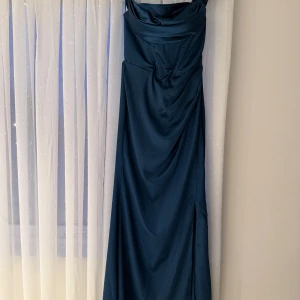 Mörkblå långklänning med draperad detalj - Elegant mörkblå långklänning med draperad överdel och smickrande passform. Klänningen har en stilren design med en subtil slits och en dragkedja i ryggen för enkel påklädning. Perfekt för en speciell kväll!